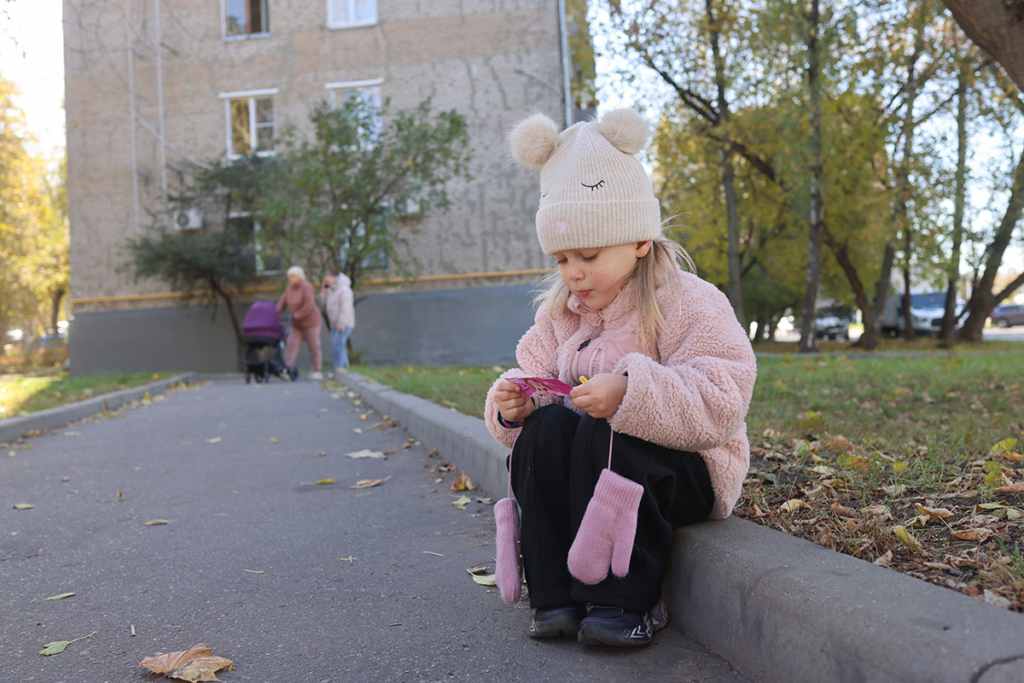 Ксения. Маленькая девочка в розовой курточке и белой шапочке на улице сидит на бордюре